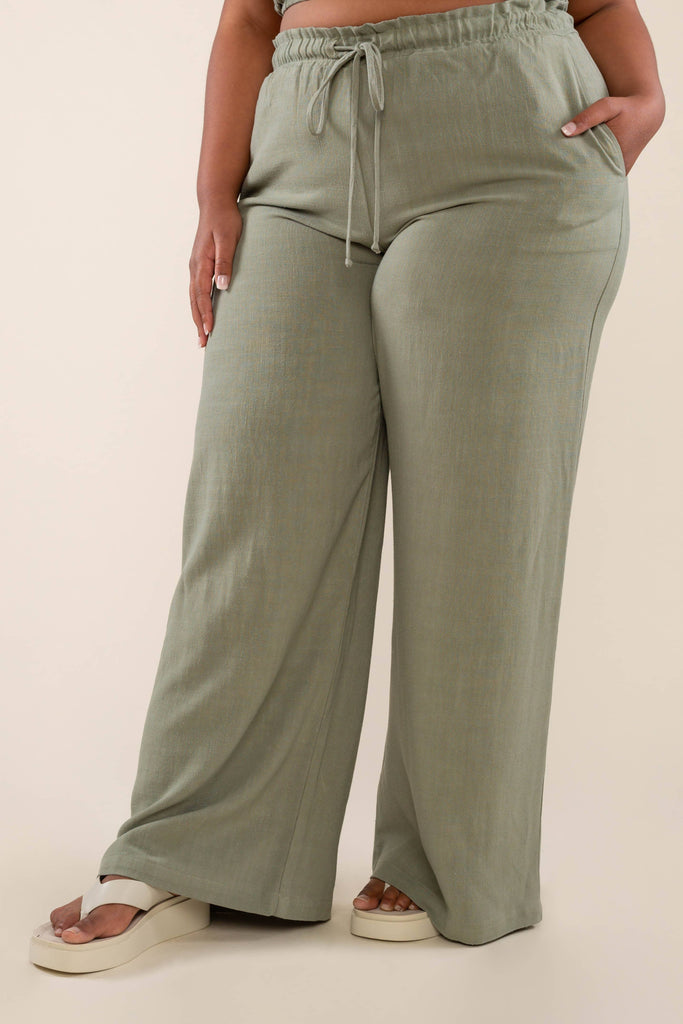 A person wearing high-waisted, loose-fitting Cove Linen Wide Leg Pant stands against a plain beige background. The wide-leg pants, made from sustainable fabric, feature an elastic waistband with a drawstring. The person is also wearing beige platform shoes and has one hand resting in a pocket.