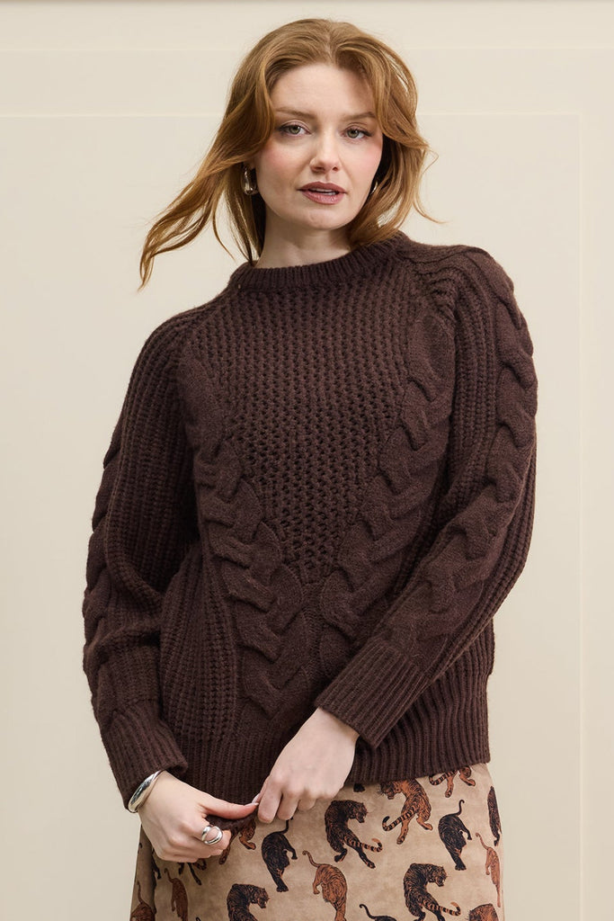 Woman wearing a brown sweater and patterned skirt standing against a beige wall.