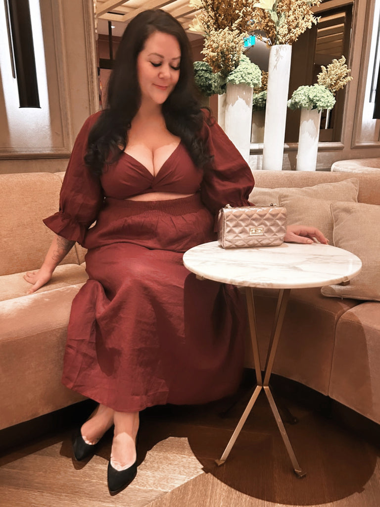 Plus size woman, MsLindsayM is seated in a hotel lobby wearing a plus size maxi dress from Laluxe Femme.