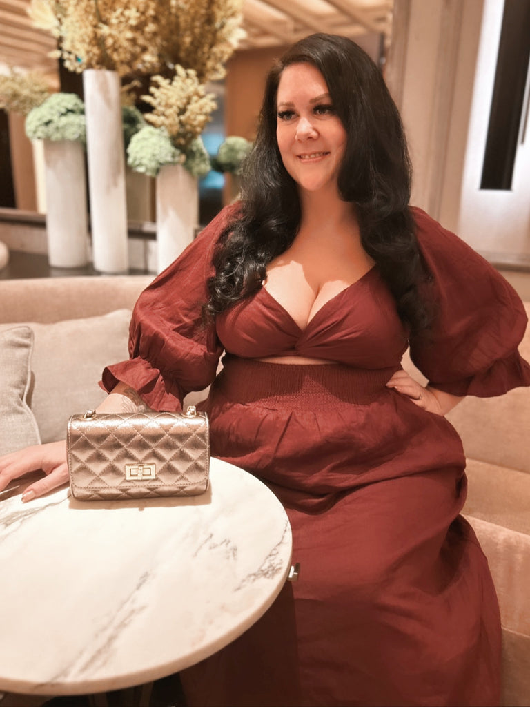 Plus size woman, MsLindsayM is seated in a hotel lobby wearing a plus size maxi dress from Laluxe Femme.